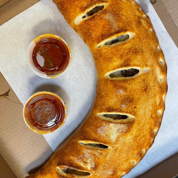 Golden-brown calzone with slits revealing filling, served with two cups of dipping sauce in a pizza box.